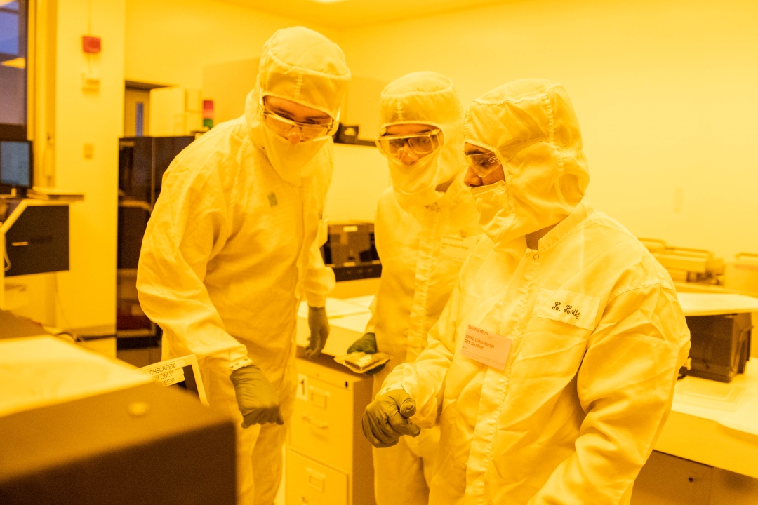 'Three people in a microchip clean room wearing clean suits, goggles, masks, and gloves. The image has a yellow tint.'